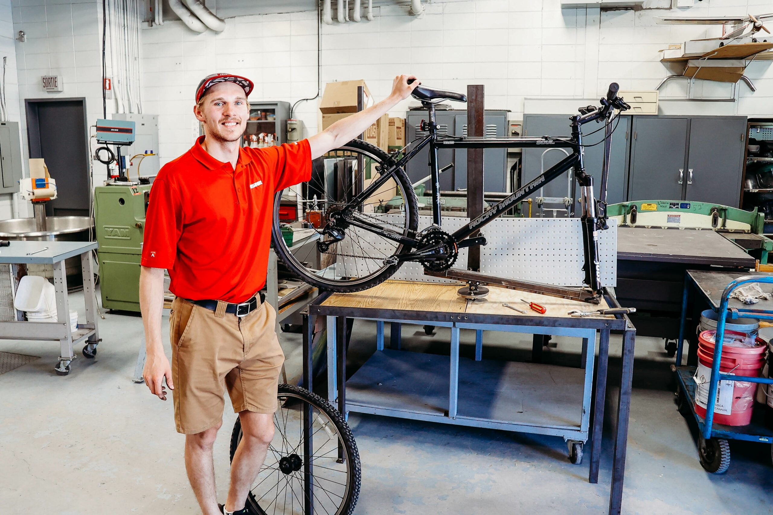 Un étudiant sourit à la caméra dans un atelier de réparation en mécanique. Un vélo est suspendu sur la table.