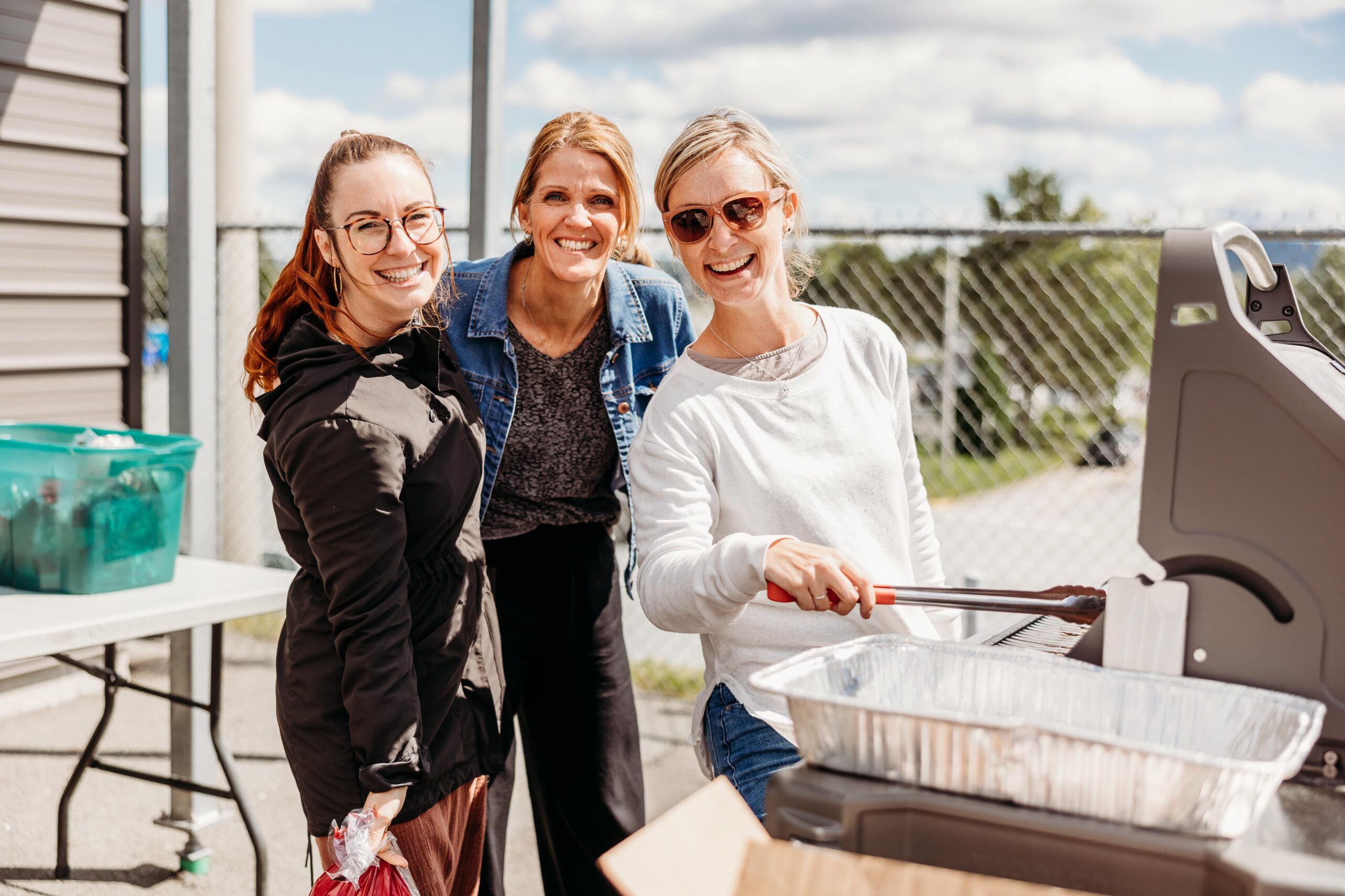 Trois femmes souriantes regardent la caméra alors qu'elles préparent de la nourriture au barbecue dans un contexte d'événement social.