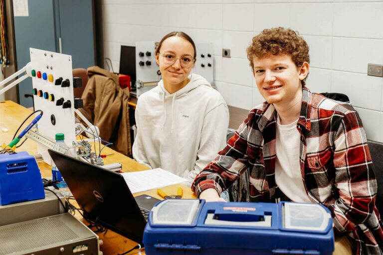 Deux étudiants sourient à la caméra assis à un poste d'électronique.