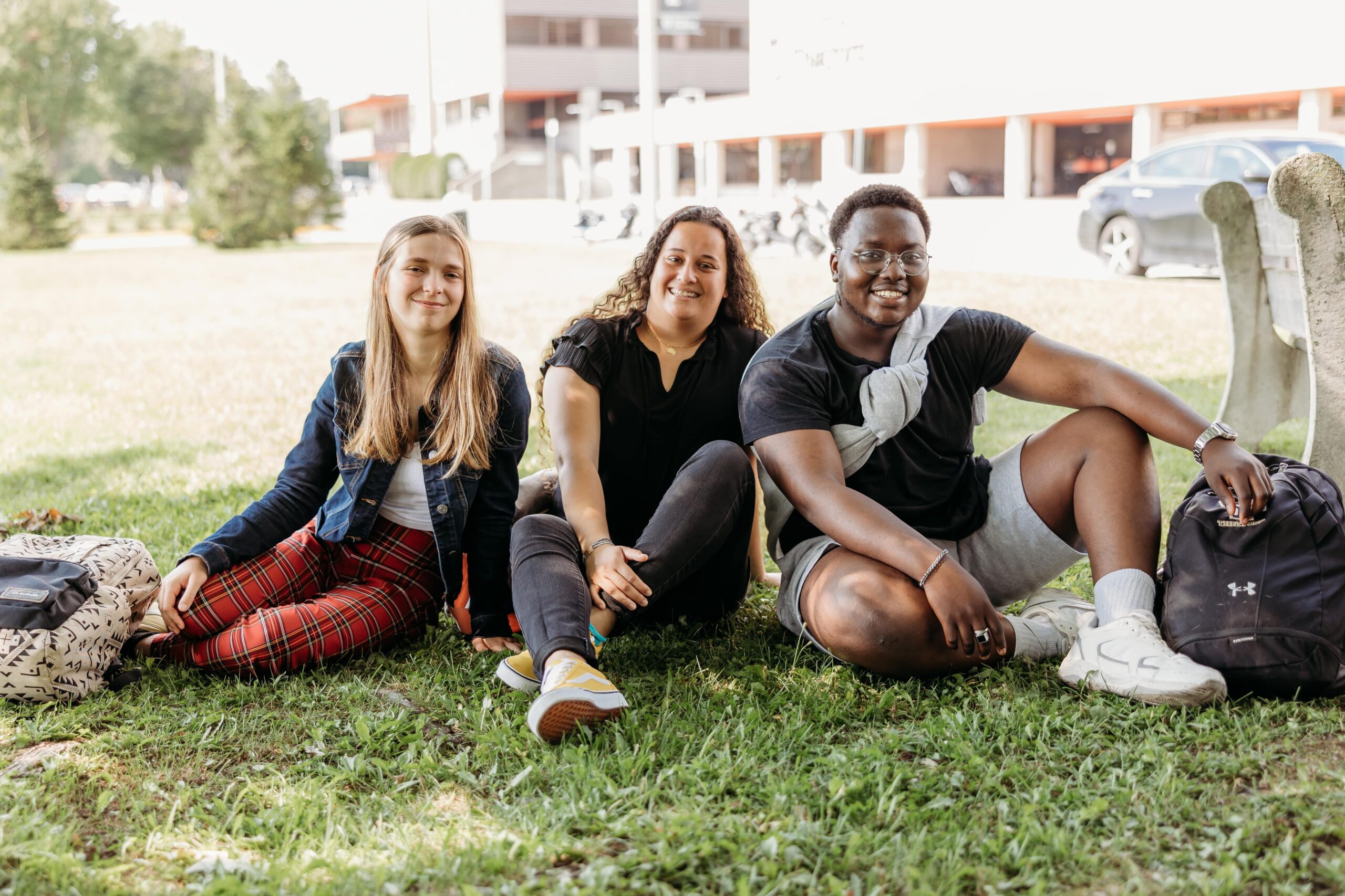 Trois étudiants assis sur le gazon sourient à la caméra.