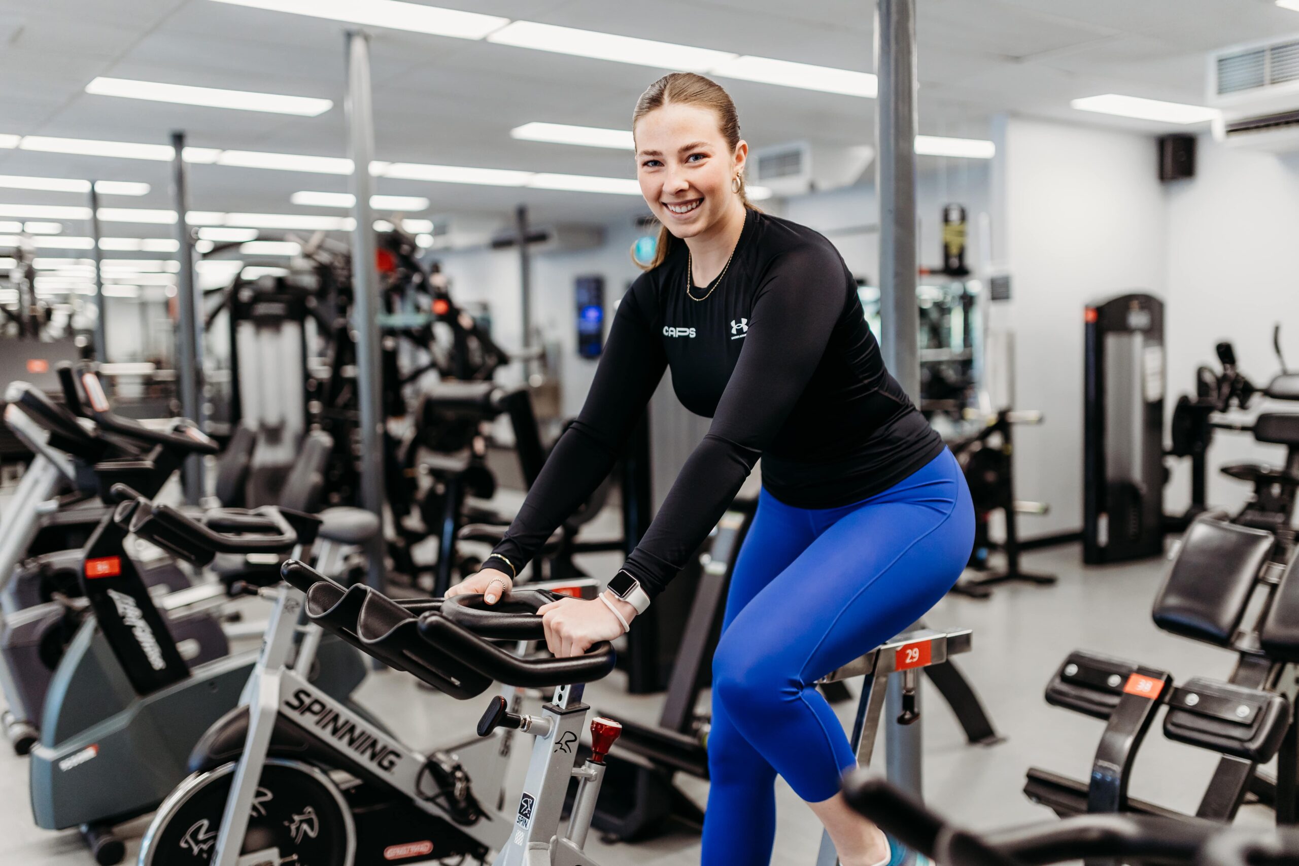 Une jeune femme sourie à la caméra alors qu'elle est sur un vélo de spinning dans une salle d'entraînement.