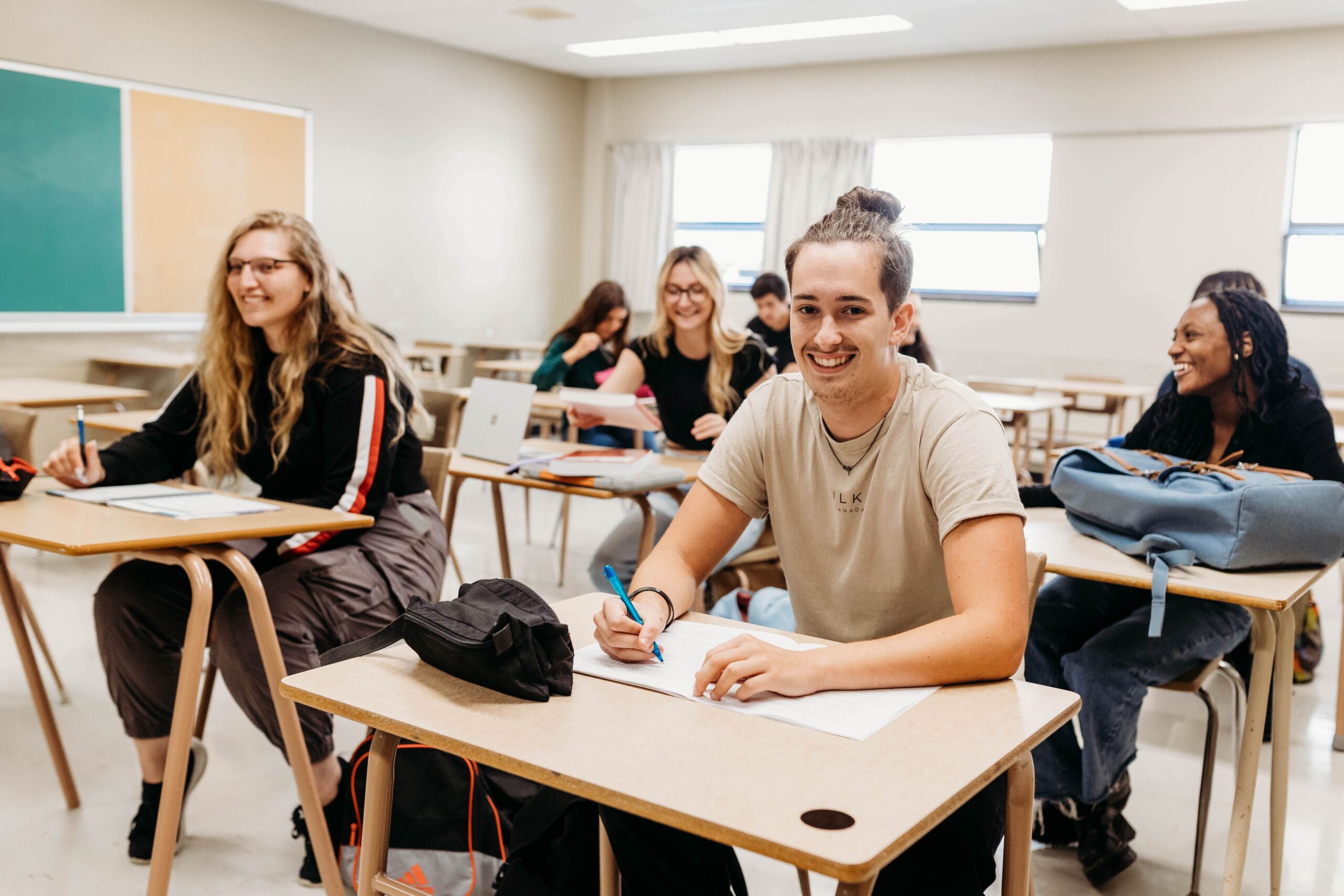Des étudiants assis dans une salle de classe ensoleillée sourient à la caméra.