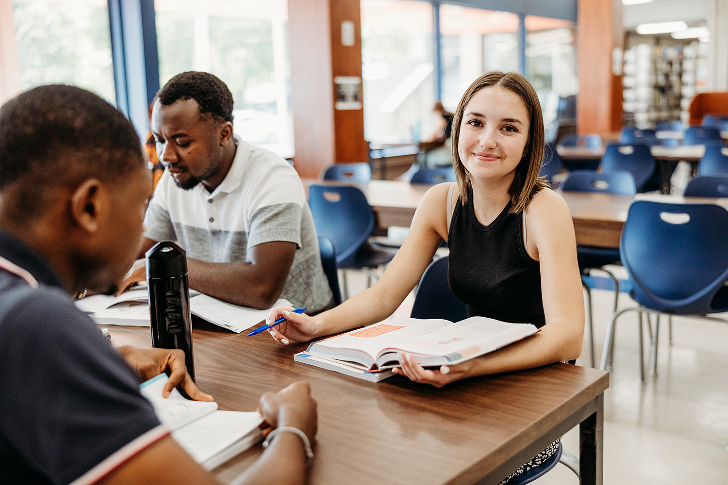 Une jeune femme sourie à la caméra accompagnée de deux collègues étudiants dans un espace commun de travail.